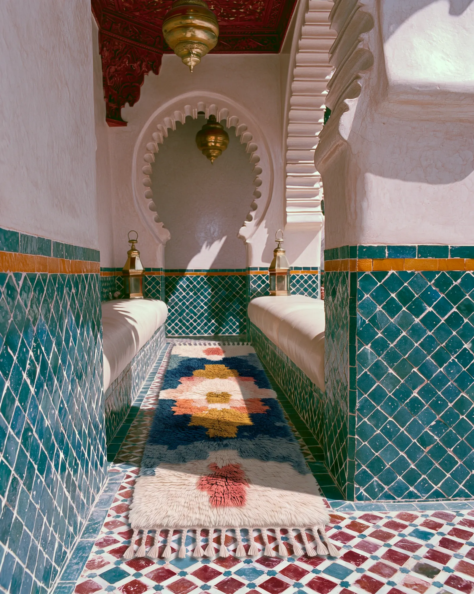 A riad salon with a teal zellige dado, a tiled floor, a Mozaik runner, brass lanterns and a carved arch: the tile geometry behind the Mozaik collection