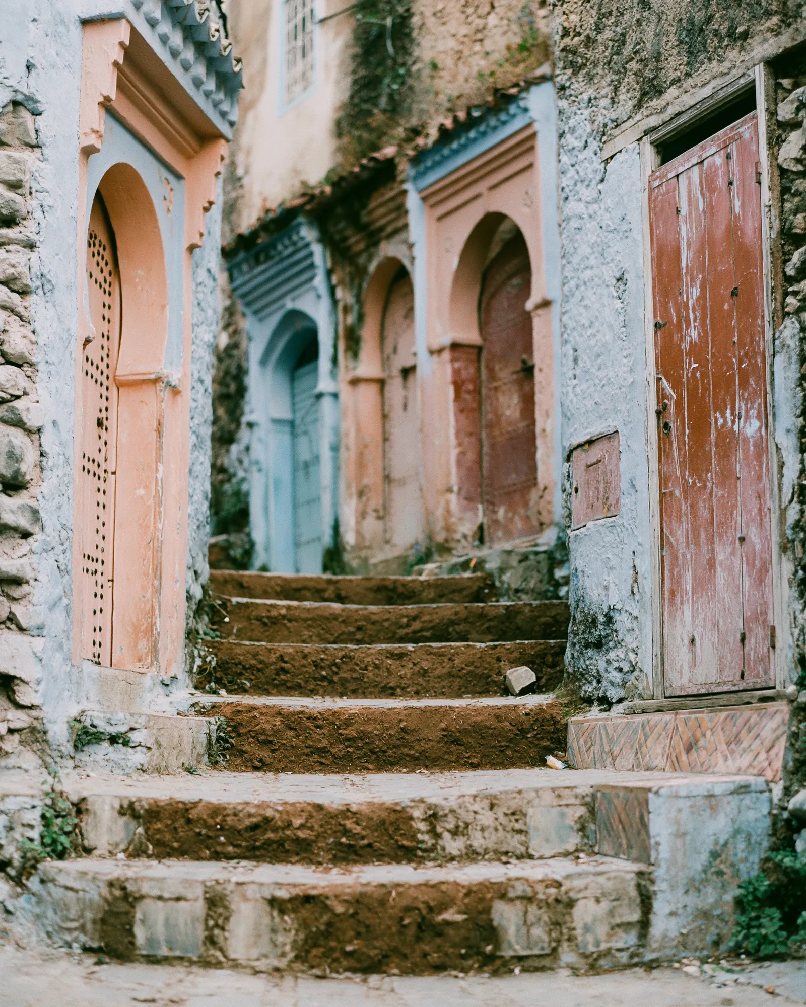 A stepped Chefchaouen alley with dusty-blue walls and peach and rust arched doors, the inspiration for the Ahidous Dusty Blue & Brown rug