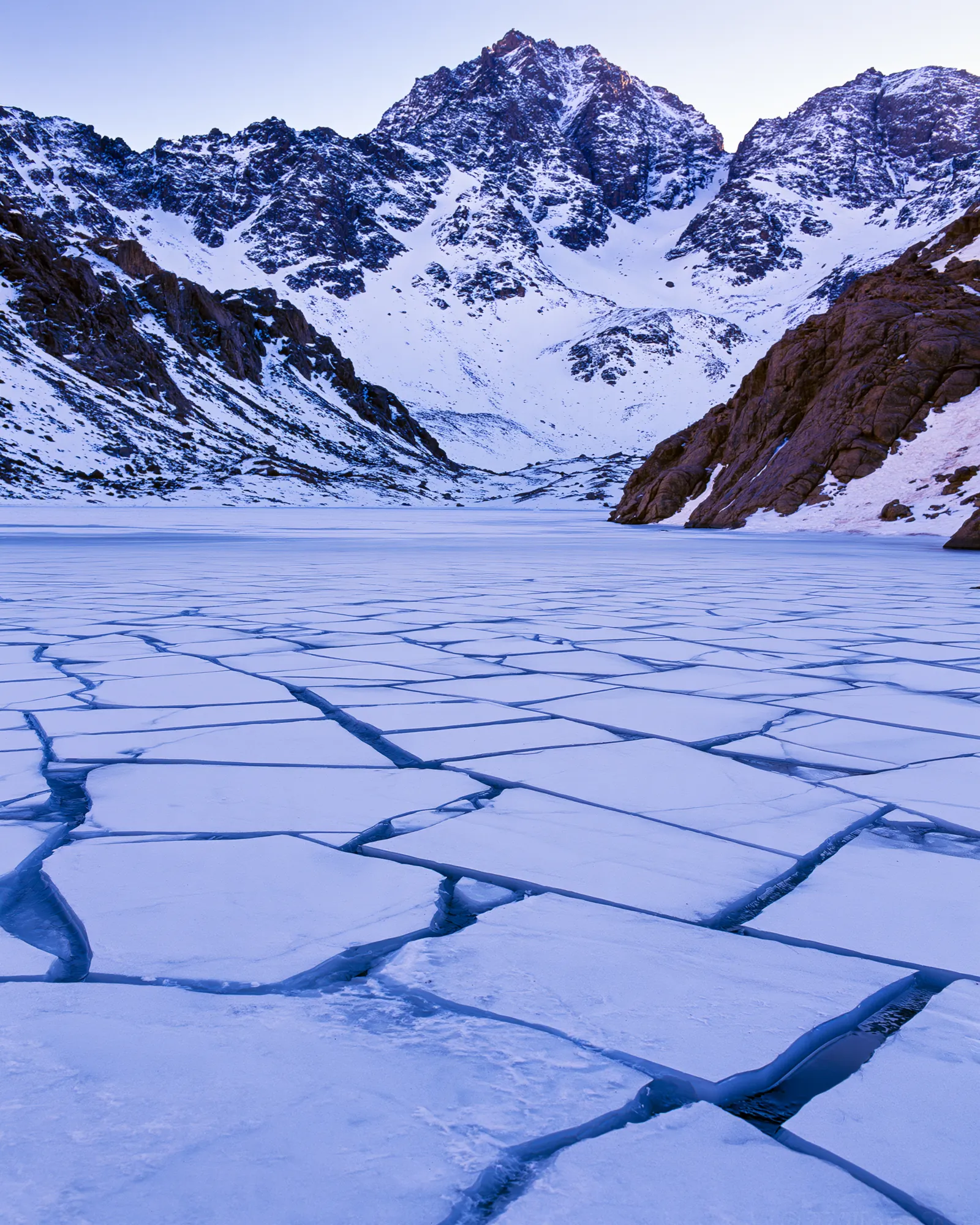 A frozen mountain lake cracked into indigo plates below snowy peaks, the inspiration for the Hamada Indigo rug