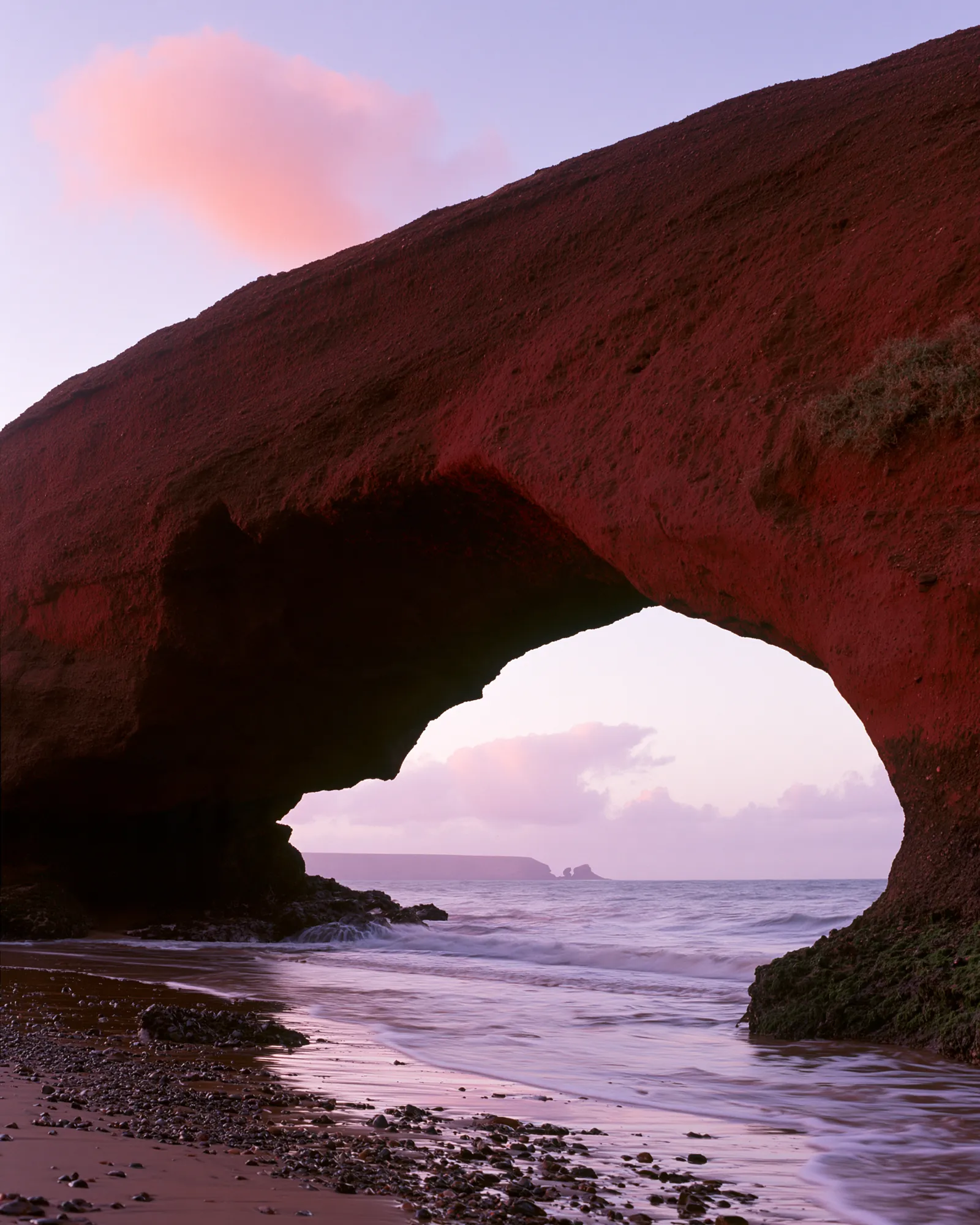 A red rock arch over the shore at Legzira in pink dusk light, the inspiration for the Legzira Burgundy & Lavender rug