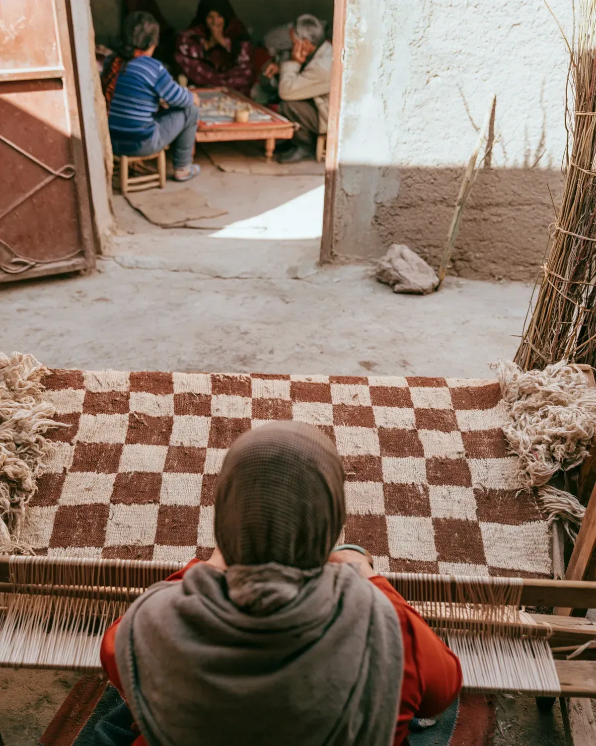 A weaver seen from behind knots a rust and ivory checkered rug at a low loom in a courtyard